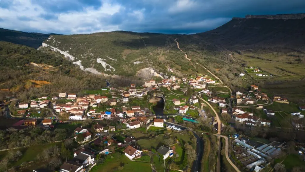 Pueblo rural entre campos y montañas Vista aérea de un pequeño pueblo entre campos y laderas boscosas bajo un cielo nublado.